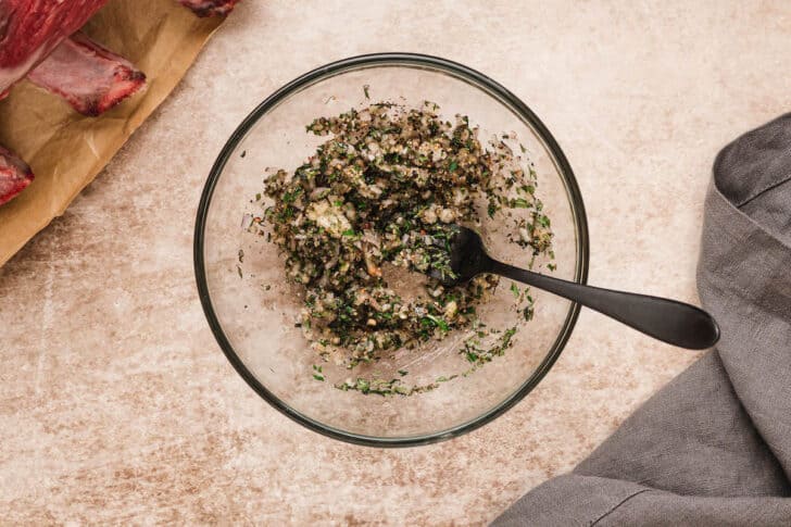 A glass bowl filled with a stirred mixture of herbs, spices, garlic and shallots.