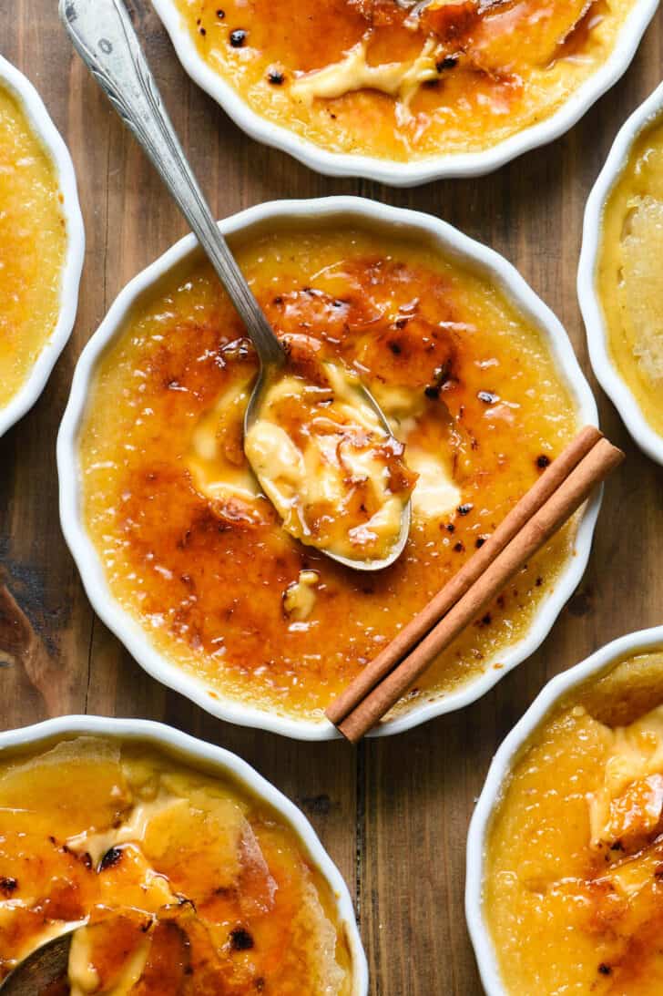 A circular white ceramic ramekin filled with an orange-hued custard topped with caramelized sugar, garnished with a cinnamon stick, with a spoon digging in.