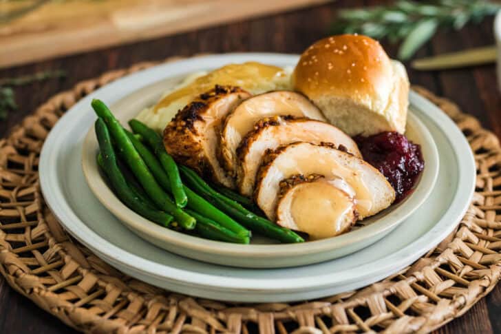 A turkey tenderloin recipe, sliced, on a plate with green beans, mashed potatoes and gravy, a roll and cranberry sauce.
