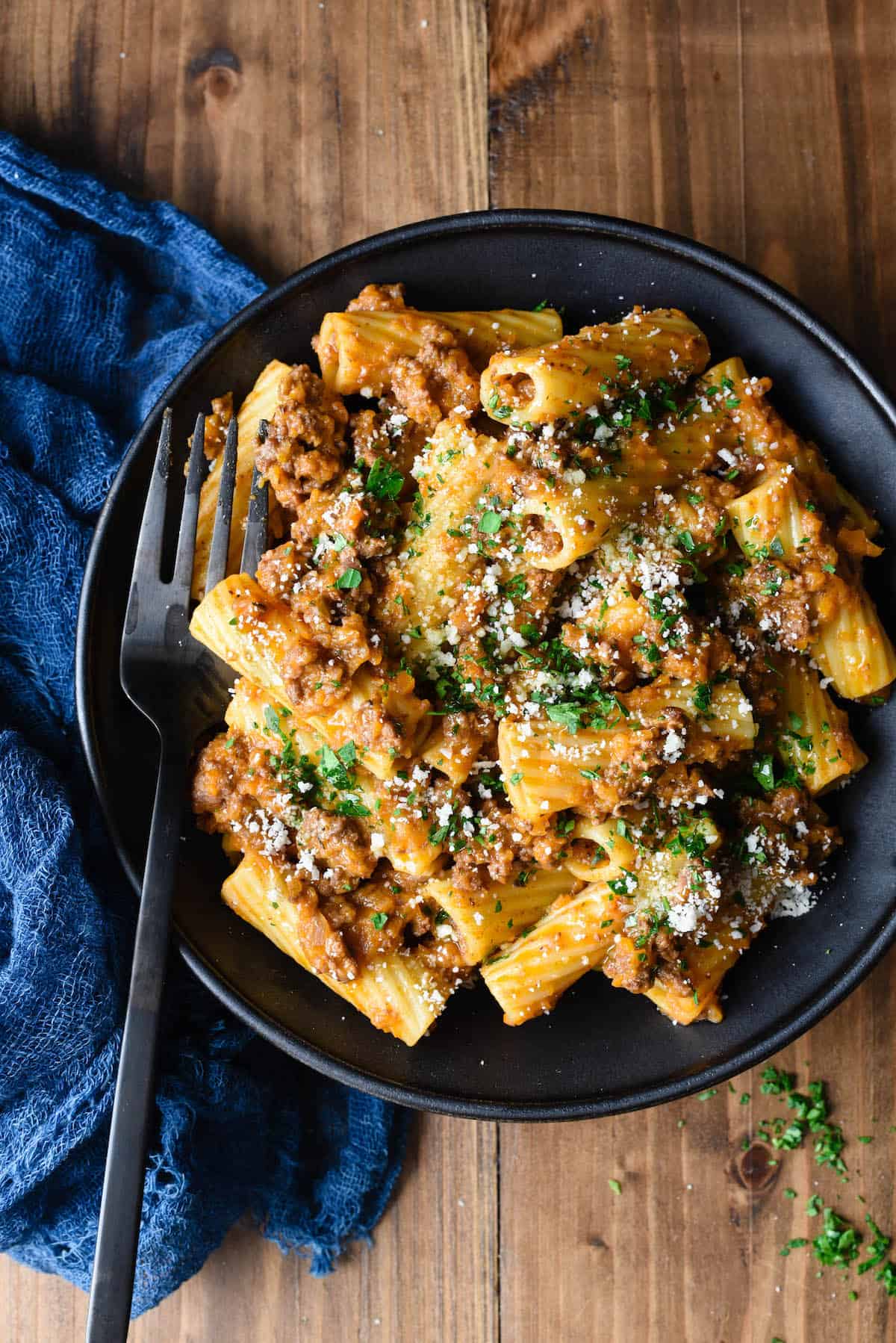 Overhead photo of rigatoni with meat sauce in black bowl, with black spoon and blue linen alongside.
