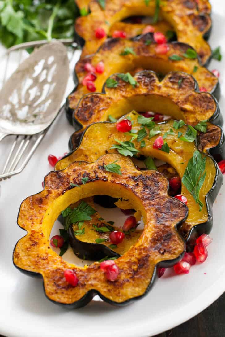 Roasted acorn squash slices garnished with pomegranate seeds and chopped parsley, lined up on a white platter.