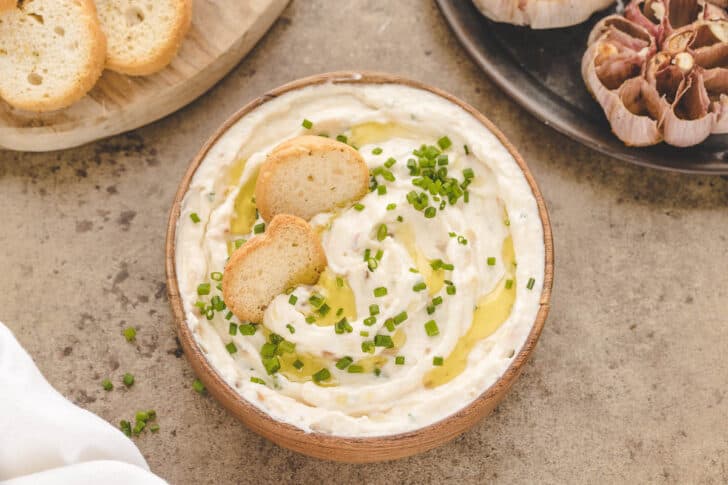 A round wooden bowl filled with roasted garlic spread, garnished with chives, toasted bread and oil.