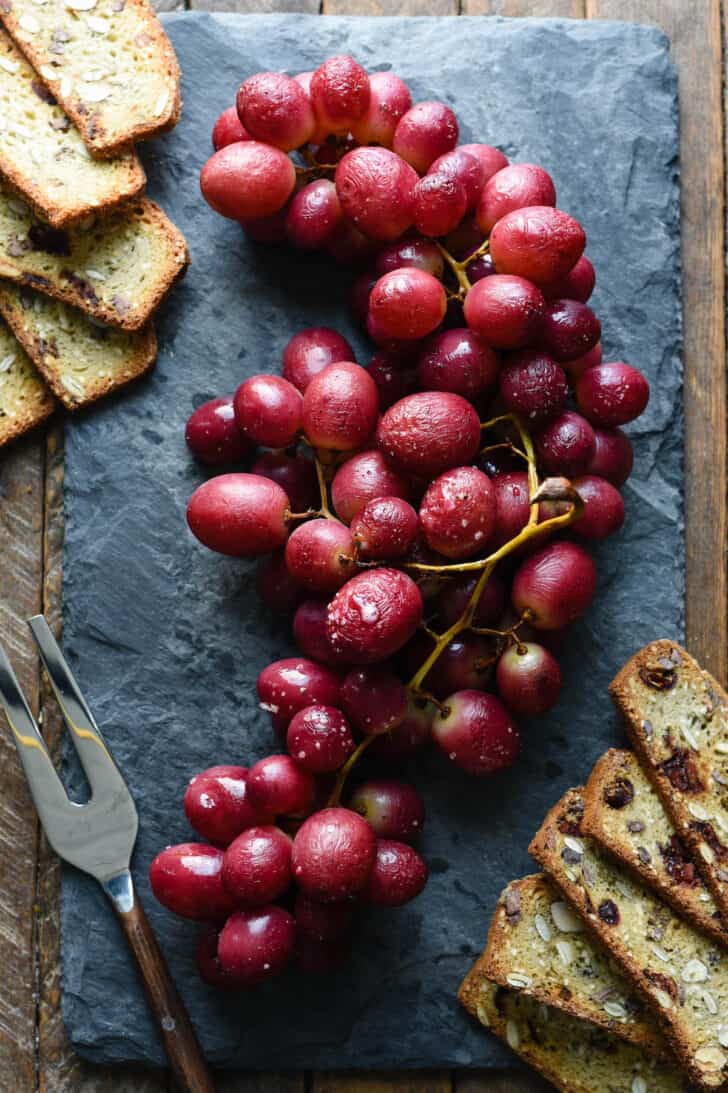 Red roasted grapes on their vines, on a slate board, with crackers and a cheese fork alongside.