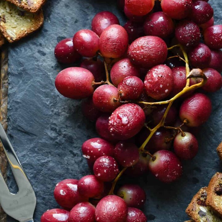 Red roasted grapes on their vines, on a slate board, with crackers and a cheese fork alongside.