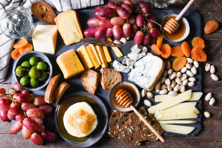 A slate cheese board filled with a variety of cheeses, pistachios, dried apricots, crackers, olives, cooked fruit and honey.