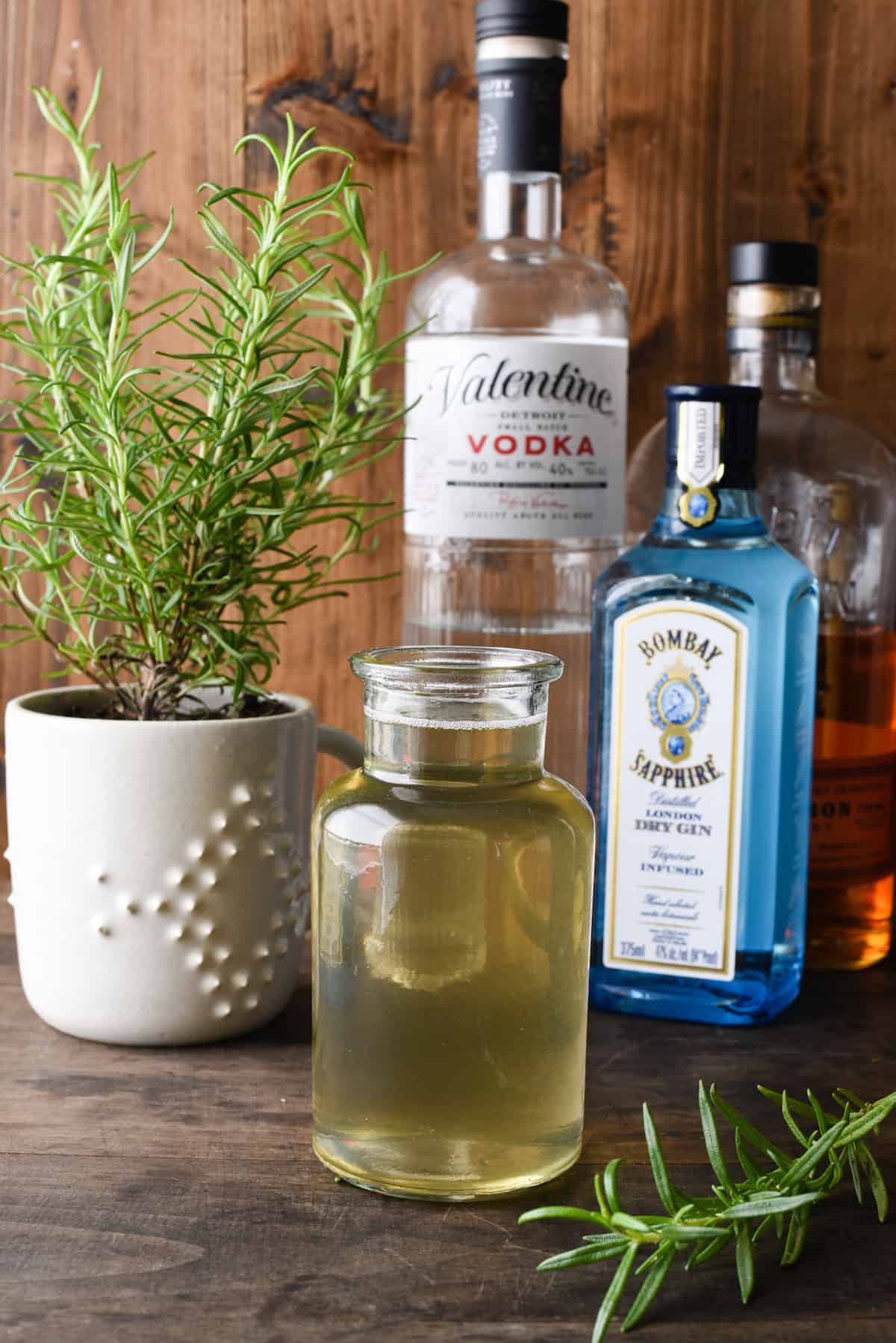 Small bottle of rosemary simple syrup with liquor bottles and rosemary plant in background.