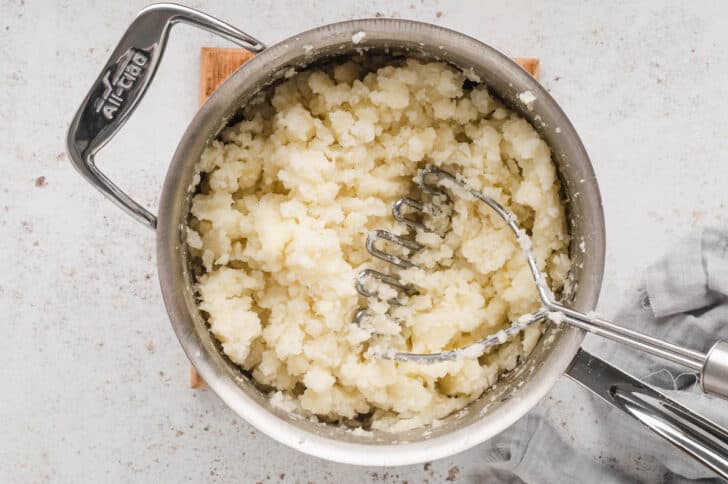 A stainless steel pot filled with potatoes that a potato masher is crushing.