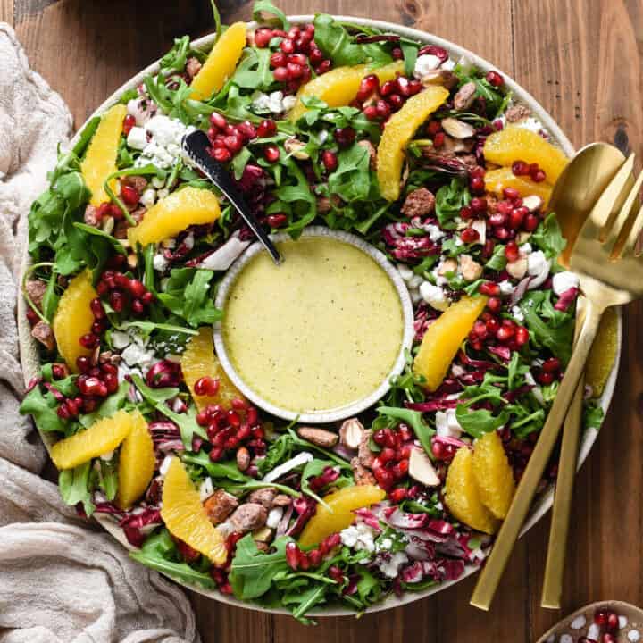 A Christmas Salad made with greens, oranges, cheese, pomegranate seeds and nuts, arranged like a wreath with a bowl of dressing in the middle.