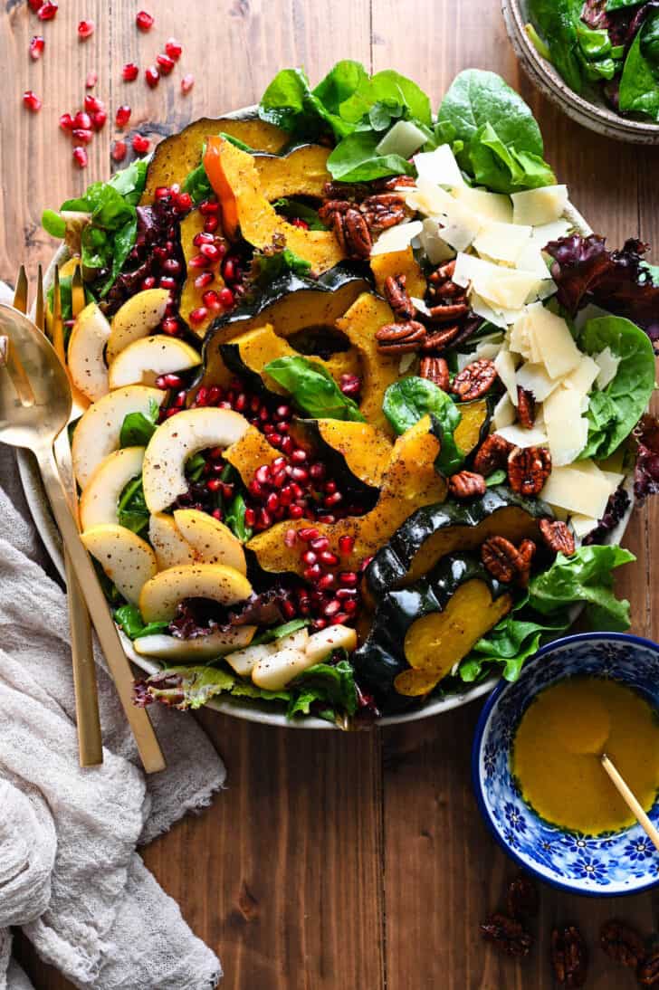 A large round platter filled with Thanksgiving Salad made with pears, pomegranate seeds, roasted acorn squash, candied pecans, Parmesan cheese and mixed greens. The toppings are arranged over the greens in rows.