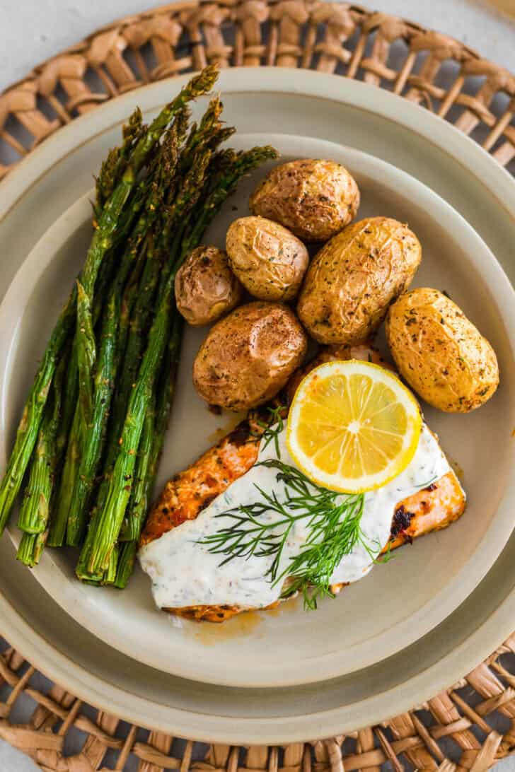 A plate of salmon, asparagus and potatoes. The fish is topped with a creamy dill sauce recipe, fresh dill, and a lemon slice.