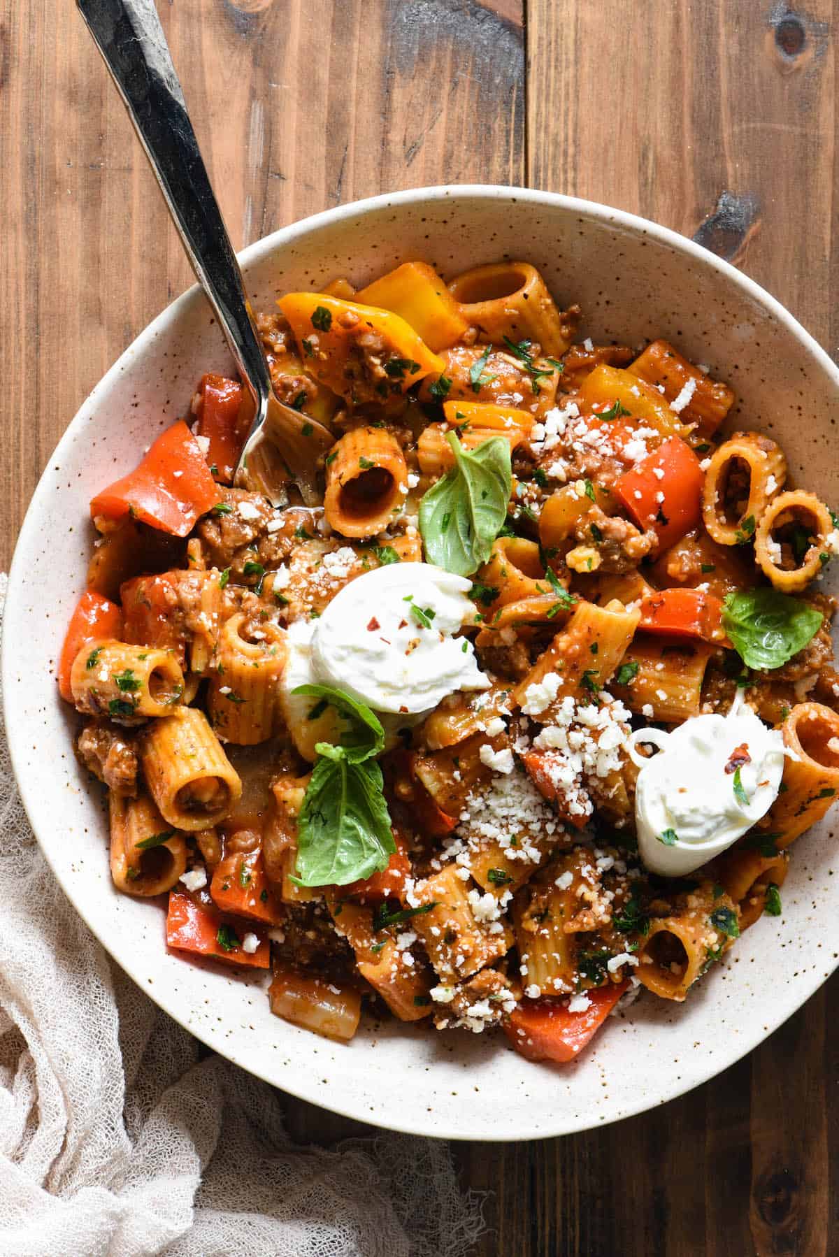 A speckled bowl filled with sausage and peppers pasta topped with fresh herbs and chunks of burrata cheese.