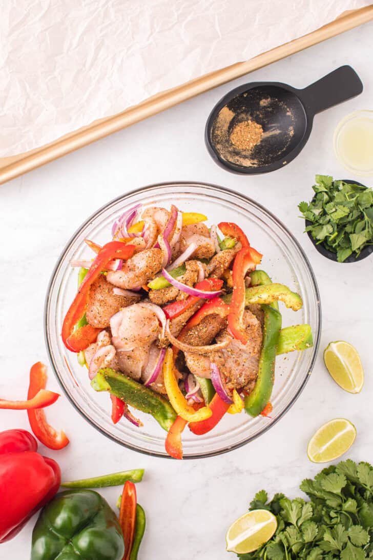 Sliced raw chicken, bell peppers and onion tossed with spices in a glass bowl.