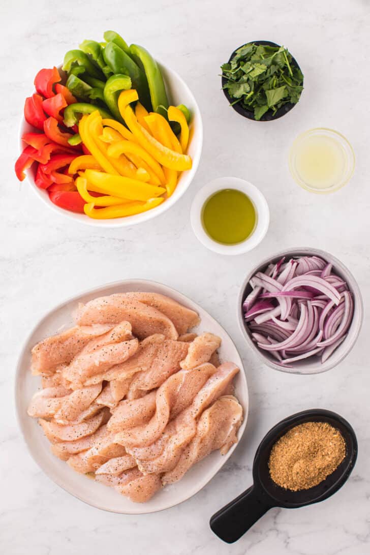 Ingredients laid out in white bowls on a light surface including sliced poultry, peppers, onions, oil and spices.