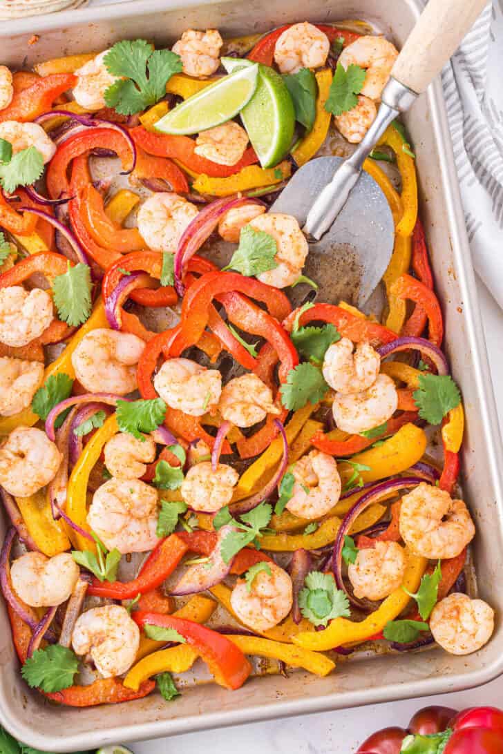 Sheet pan shrimp fajitas sprinkled with cilantro on a light marble surface, with a metal spatula scooping some off the pan.