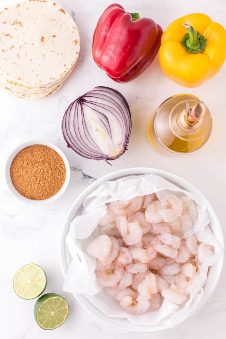 All of the ingredients needed for a shrimp fajita recipe, including shrimp, bell peppers, onion, spices, tortillas, oil and limes.