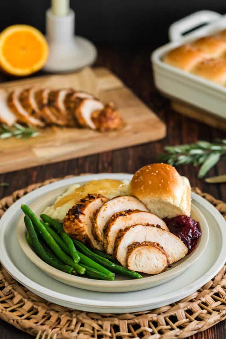 A turkey tenderloin recipe, sliced, on a plate with green beans, mashed potatoes and gravy, a roll and cranberry sauce.