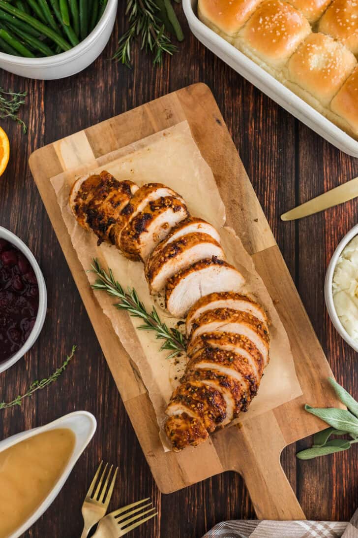 Sliced turkey breast tenderloin recipe on a wooden cutting board, surrounded by side dishes.