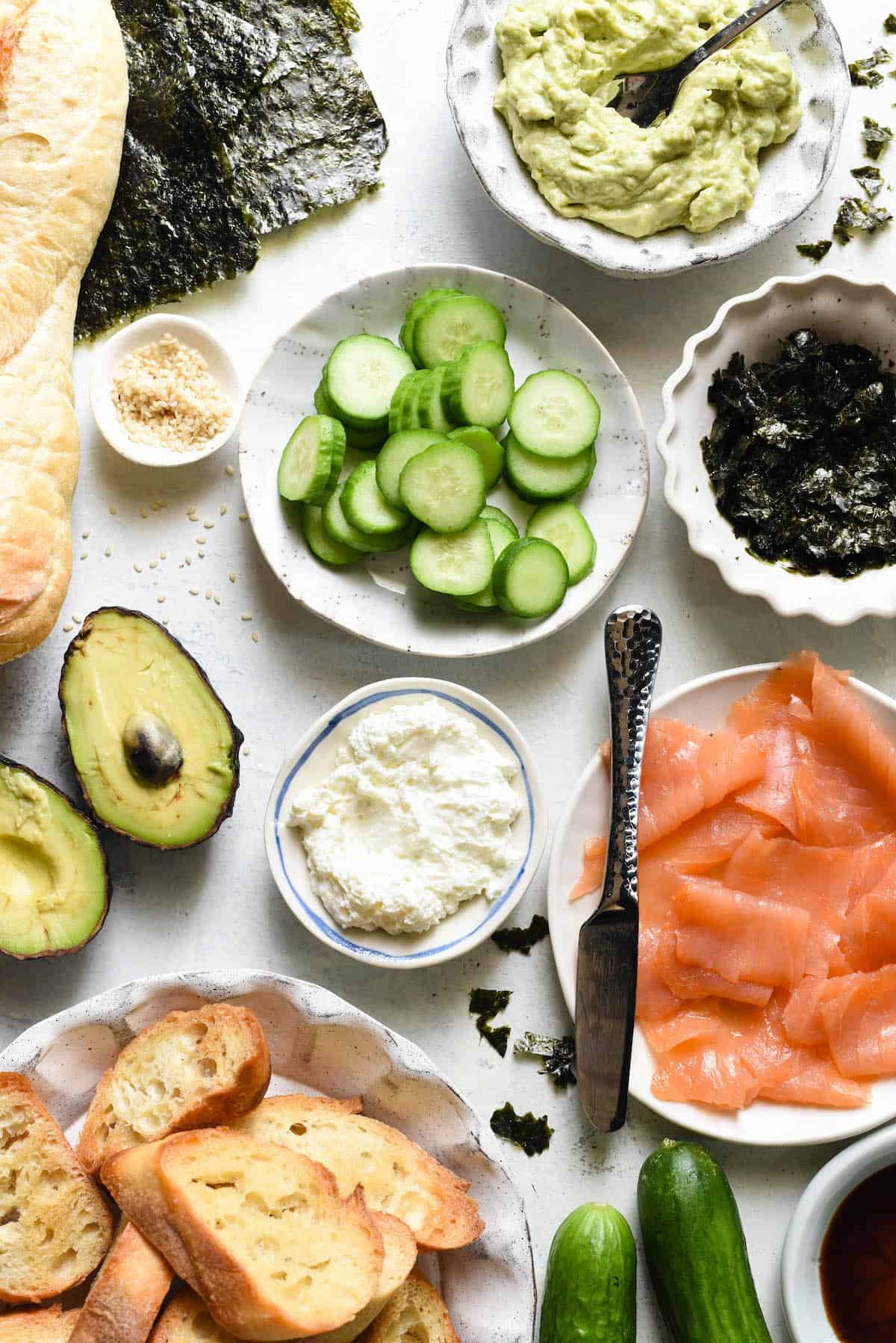 Ingredients for salmon bruschetta arranged in small bowls on white tabletop. Avocado spread, sliced cucumber, seaweed, smoked salmon, cream cheese, sesame seeds, sliced baguette.