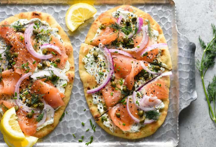 Flatbread pizzas on a metal textured tray made with naan bread, cream cheese, herbs, everything bagel seasoning, lox, capers and red onion.