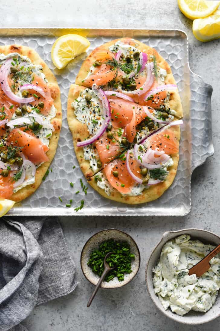 Smoked salmon pizzas on a metal textured tray made with naan bread, cream cheese, herbs, everything bagel seasoning, lox, capers and red onion.