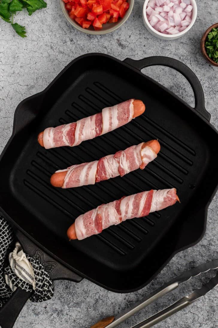 Three bacon wrapped hot dogs on a square black grill pan.