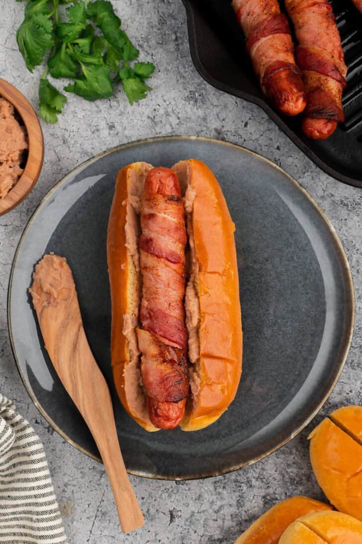A bacon wrapped hot dog in a bun spread with refried beans on a gray plate.