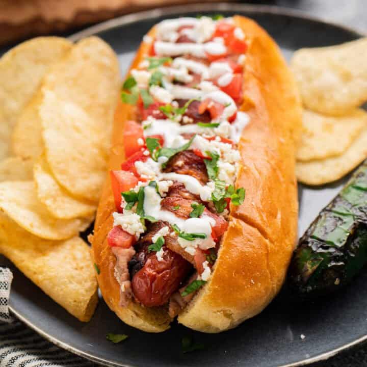 Bacon wrapped hot dog topped with sour cream, tomatoes, cilantro and cheese, on a black plate with potato chips.