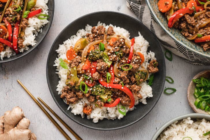 A black bowl filled with white rice topped with ground beef stir fry and veggies, garnished with green onions and sesame seeds.