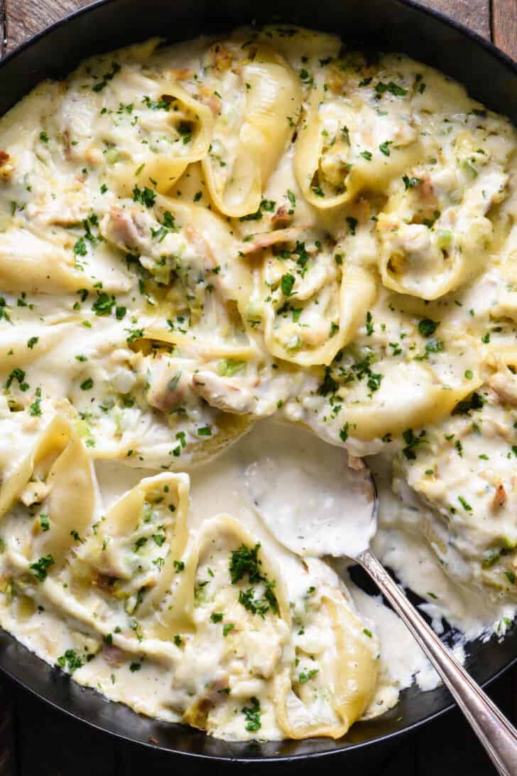 A cast iron skillet filled with alfredo stuffed shells, with a spoon scooping some sauce out.
