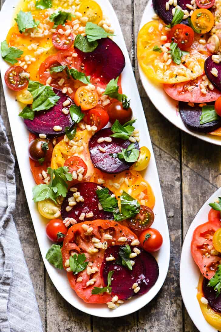 A summer beet salad recipe on an oblong white platter, with tomatoes, garnished with herbs and barley.