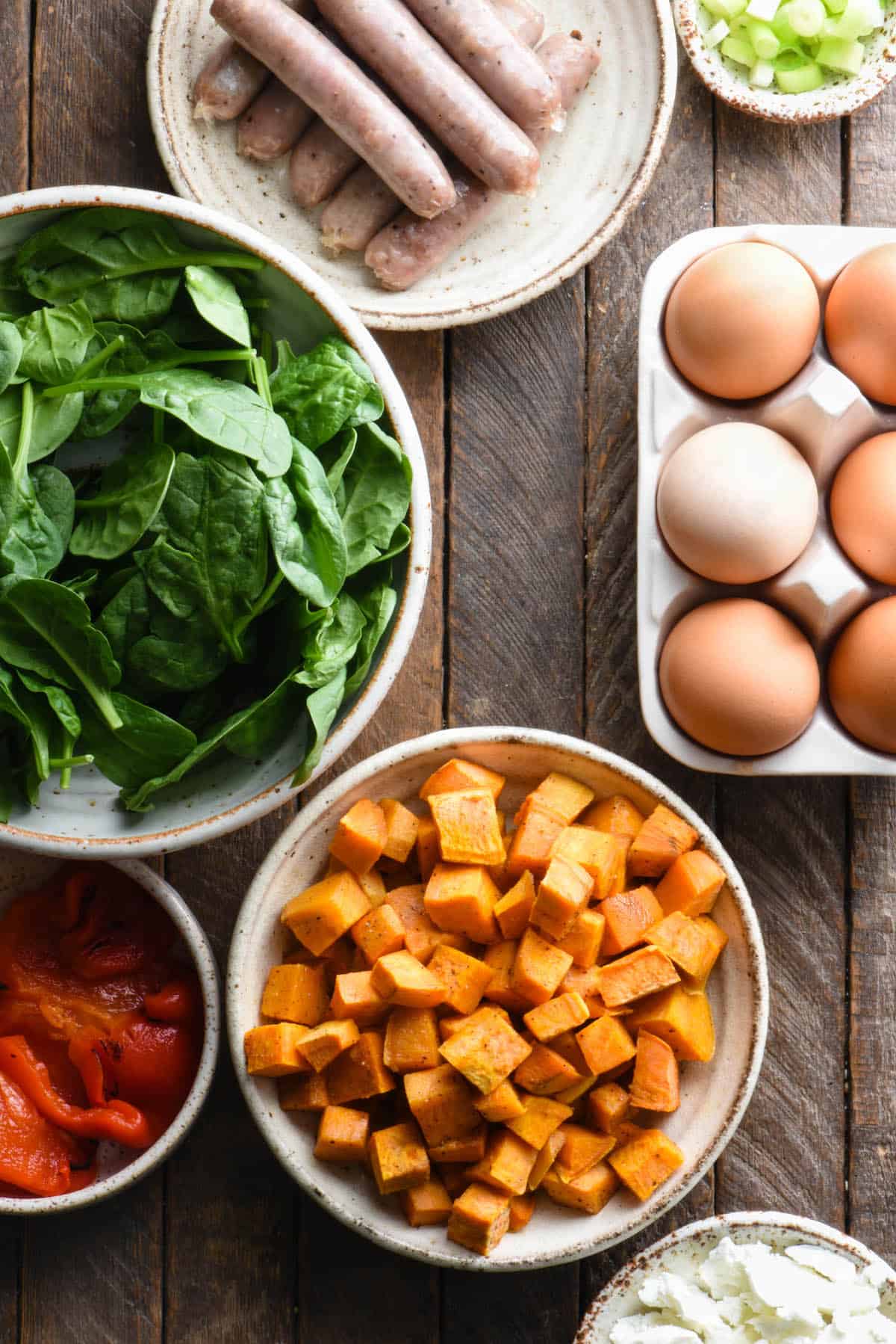 The ingredients needed for a sweet potato breakfast recipe laid out on a wooden surface, including brown eggs, spinach, sausage, sweet potatoes, cheese and red peppers.