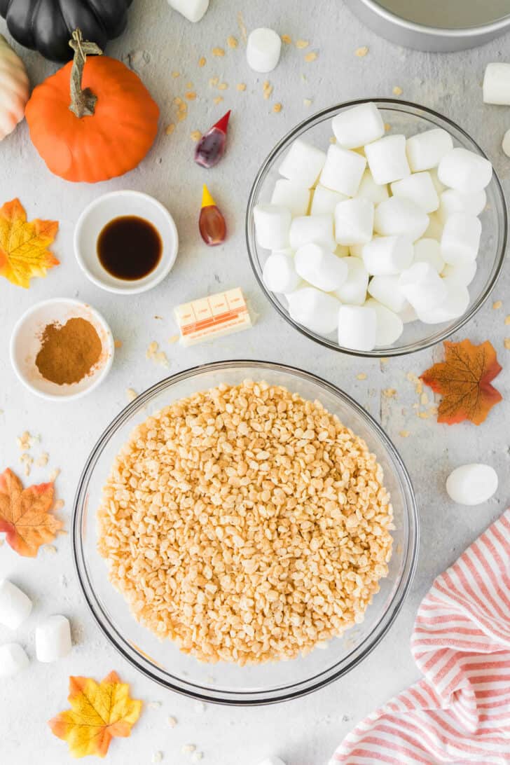 Ingredients needed for pumpkin spice Rice Krispies treats, including cereal, marshmallows, food coloring, extract and butter, laid out on a light surface.