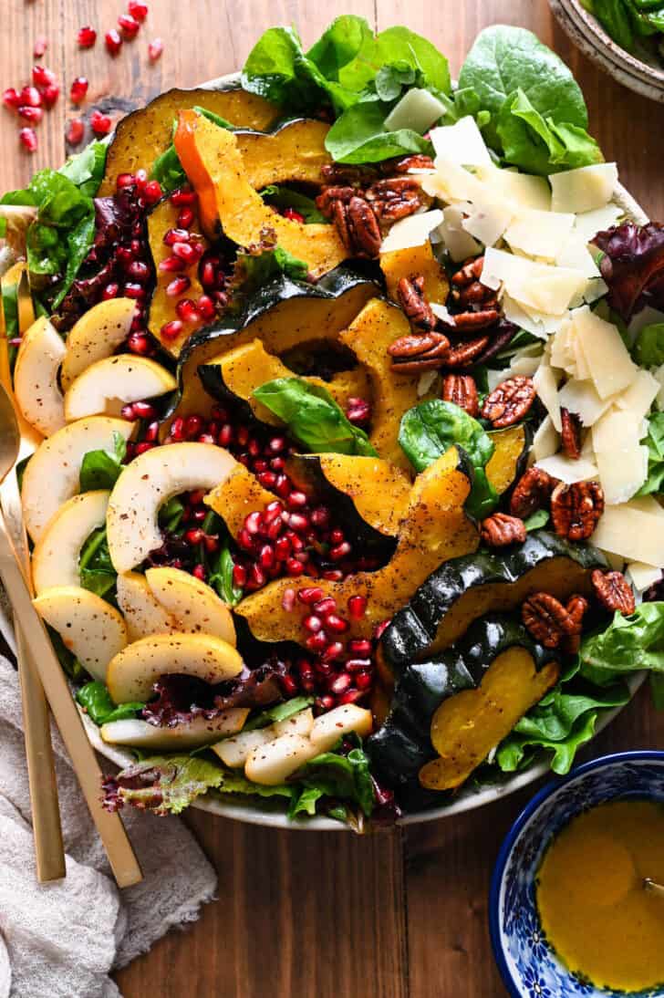 A large round platter filled with Thanksgiving Salad made with pears, pomegranate seeds, roasted acorn squash, candied pecans, Parmesan cheese and mixed greens. The toppings are arranged over the greens in rows.