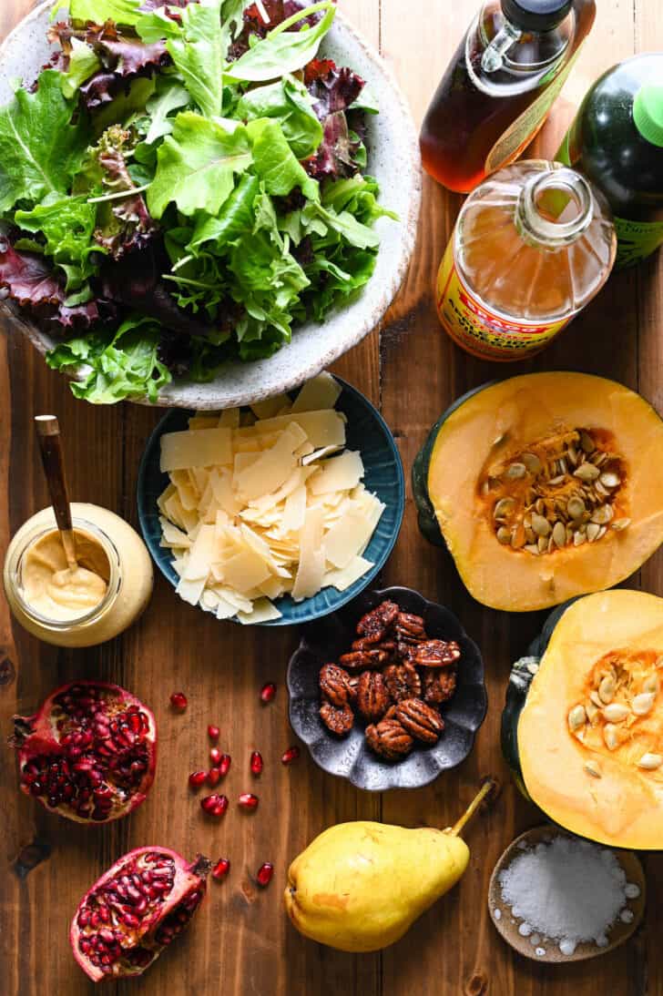 Ingredients on a wooden table including a large bowl of mixed greens, shaved Parmesan cheese, a halved acorn squash, candied pecans, a split Pomegranate, a pear, Dijon mustard and spices.