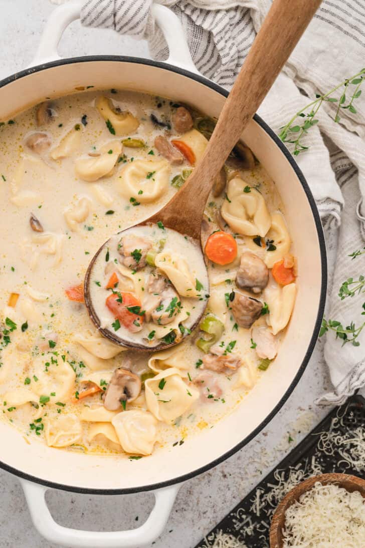 A white Dutch oven with a wooden ladle spooning out chicken tortellini soup.