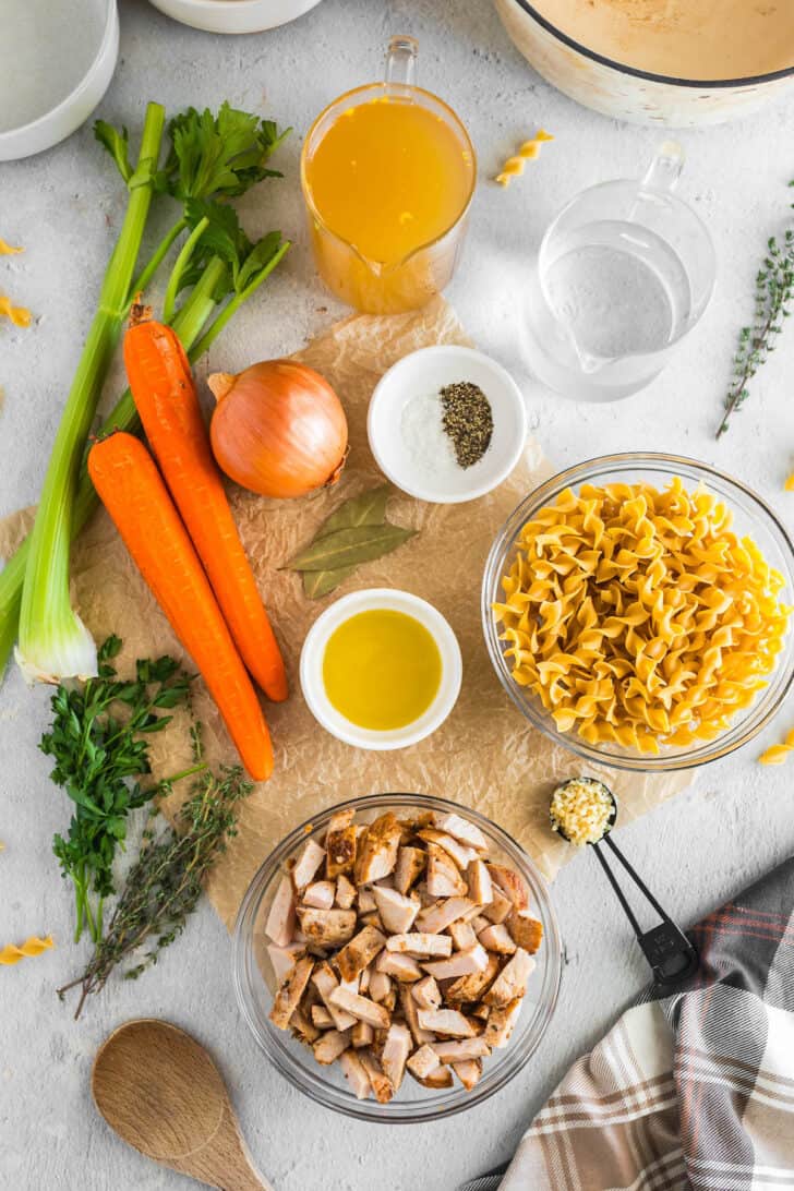 Ingredients laid out on a light surface, including carrots, celery, onion, garlic, herbs, spices, poultry and noodles.