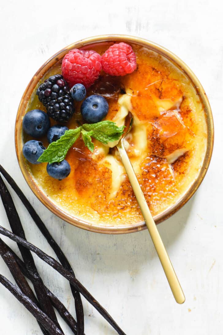 Shallow dish of vanilla creme brulee garnished with berries and mint, with a gold spoon digging in, with vanilla beans alongside.
