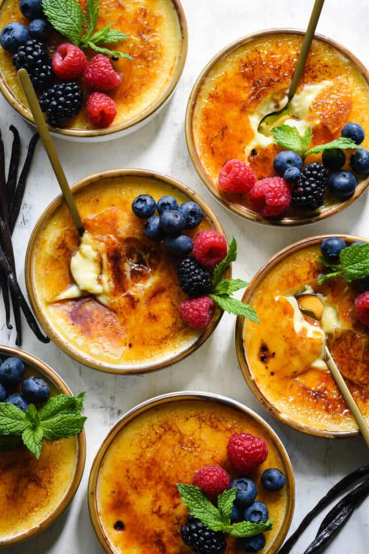 Six dishes of vanilla bean creme brulee garnished with berries and mint sprigs, on a light surface, with gold spoons digging into three of the dishes, and extra vanilla beans alongside.