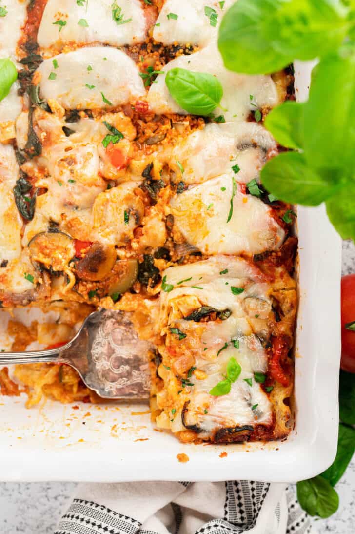 A white baking dish of veggie lasagna, with a serving utensil about to lift one piece out.