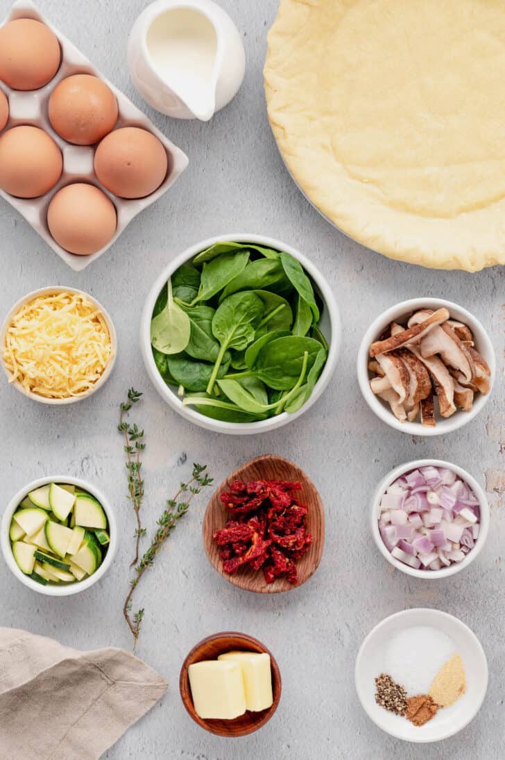 The ingredients needed for a vegetarian quiche recipe laid out on a light surface, including pie crust, eggs, veggies, cheese and herbs.