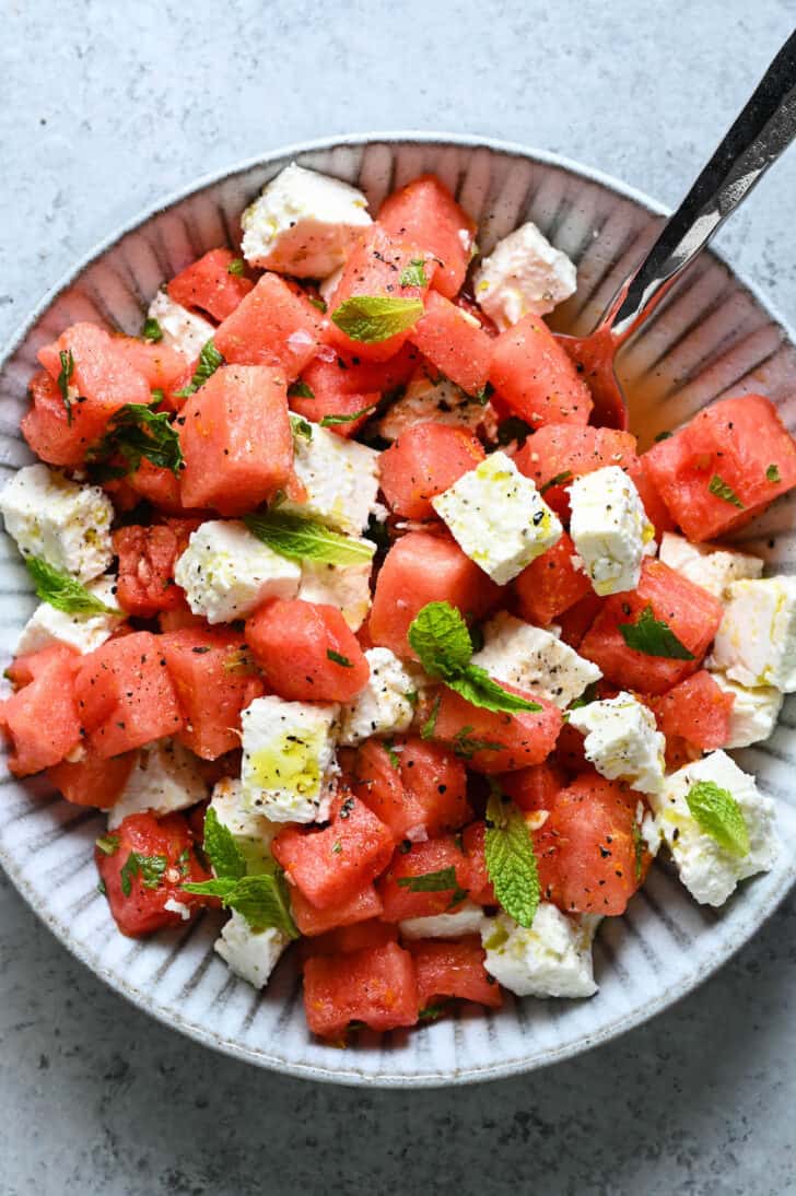 A textured gray bowl filled with watermelon feta mint salad, with a spoon digging into it.