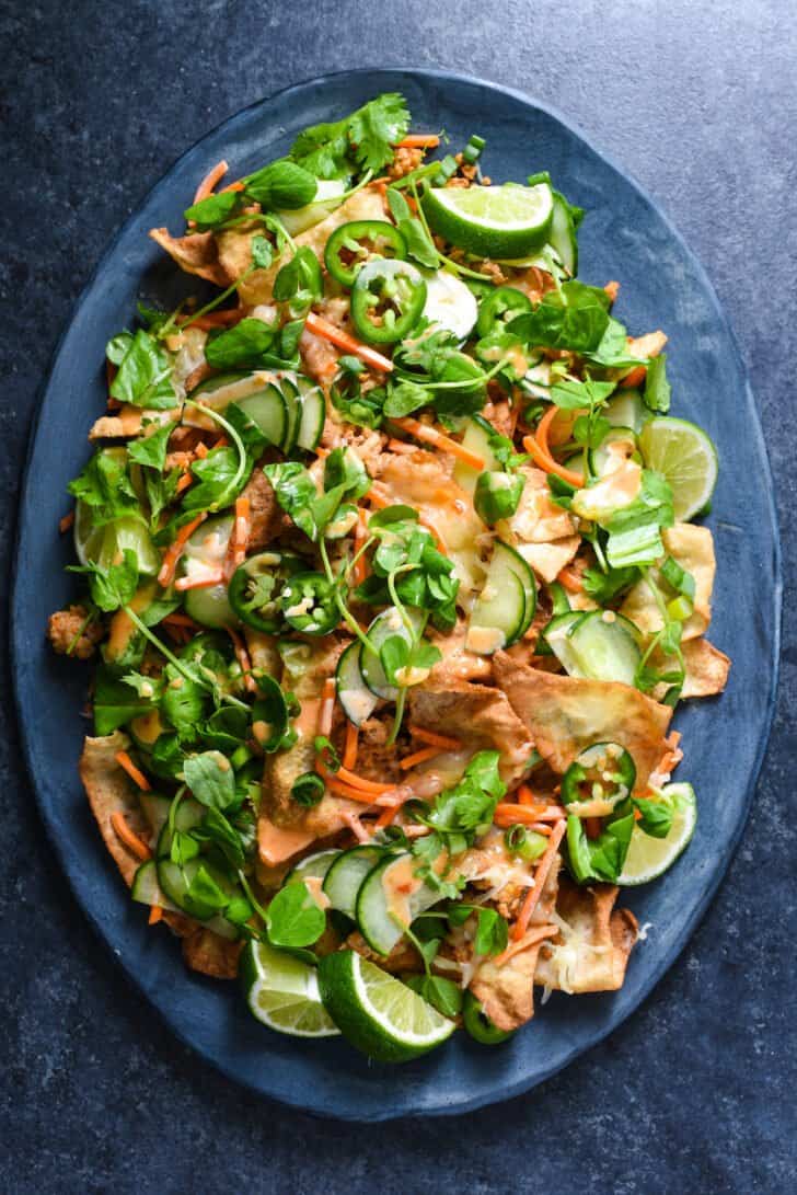 A large oval platter filled with wonton nachos made from fried wonton chips, pork, pickled veggies, cheese, herbs and spicy mayo.